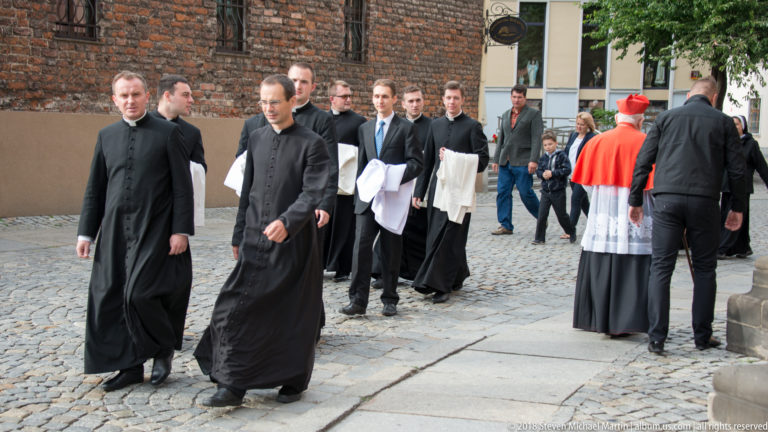 Group of Catholic priests and a senior cleric in red and white vestments walking along a cobbled street.