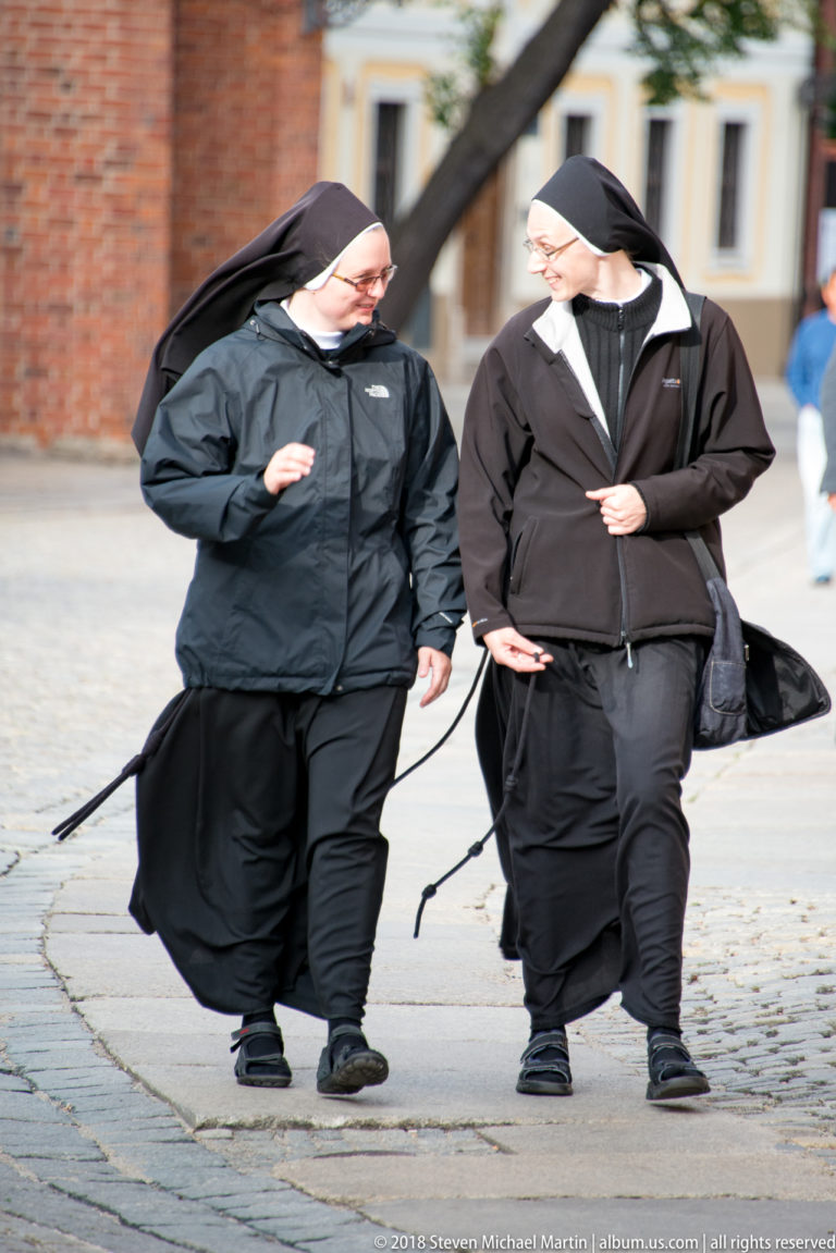 Two Catholic nuns in black-and-white habits walk together across a cobbled street.