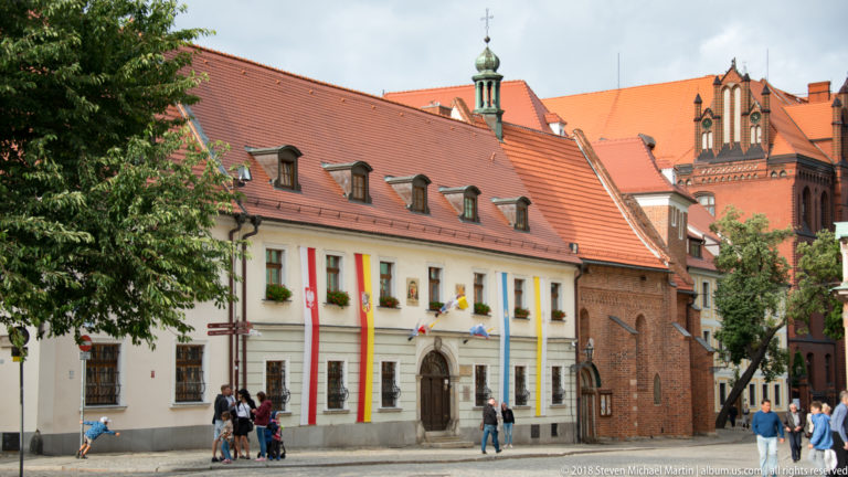 Pale historic building with flower boxes beside red-brick church structures and steep orange tiled roofs.