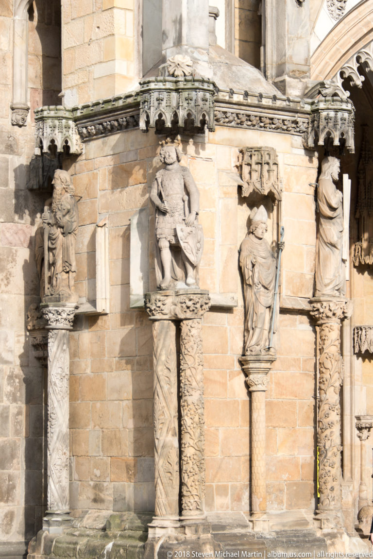 Weathered stone figures on carved columns beneath ornate capitals along a historic church exterior.