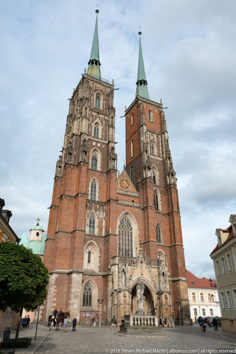 Front view of Wrocław Cathedral with twin brick towers, green spires, Gothic portals, and large central window.