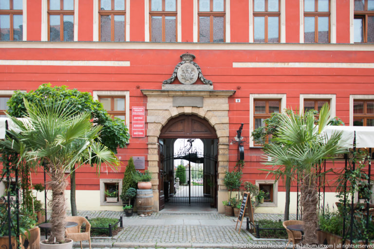 Stone-framed arched gateway in a red building, flanked by potted palms and topped by a sculpted crest.