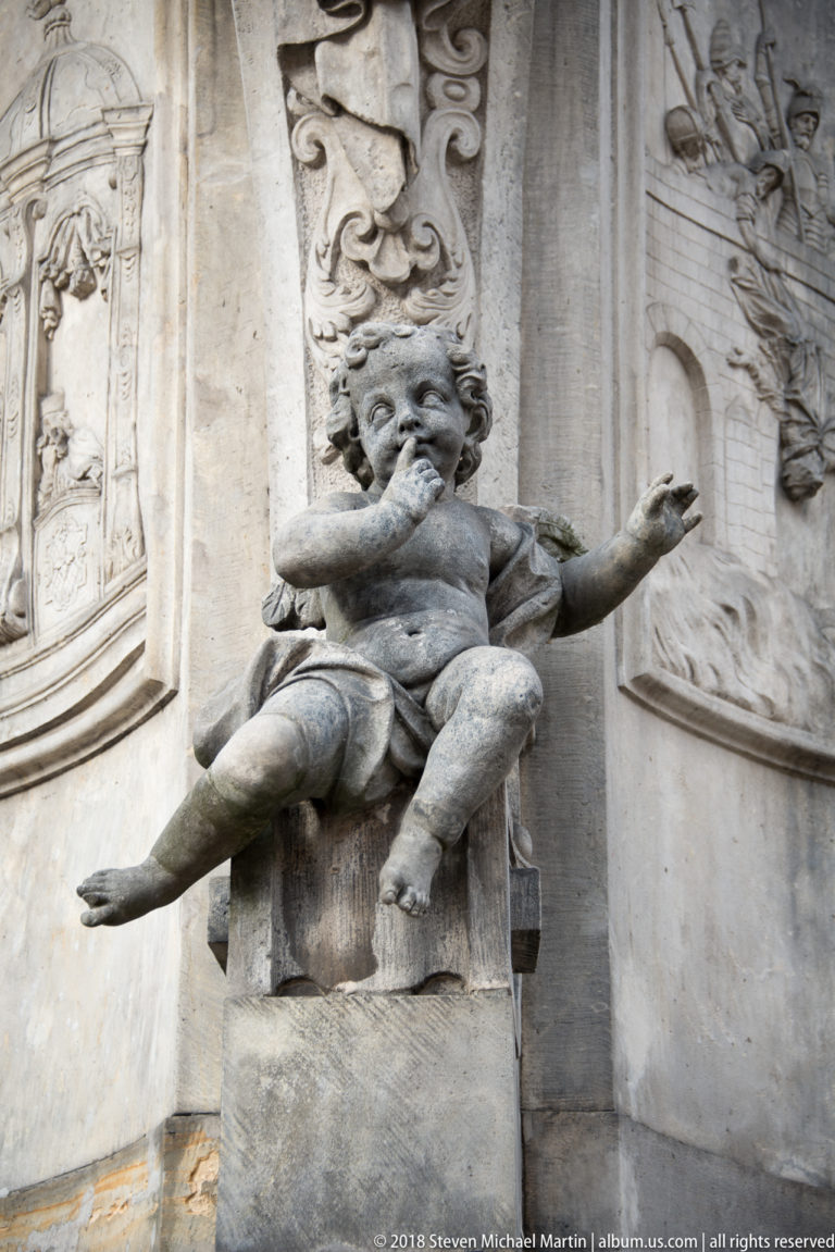 Carved childlike grotesque figure projects from an ornate stone façade surrounded by relief panels and scrollwork.