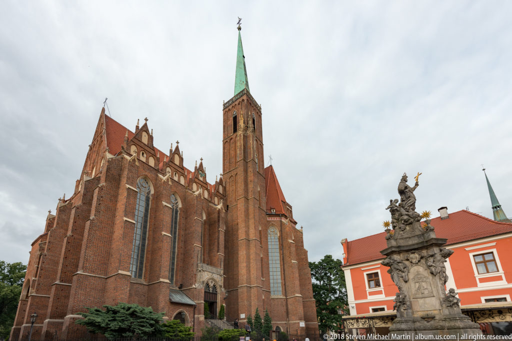 Large Gothic brick church with pointed windows, buttresses, square tower, green spire, and stone monument in foreground.