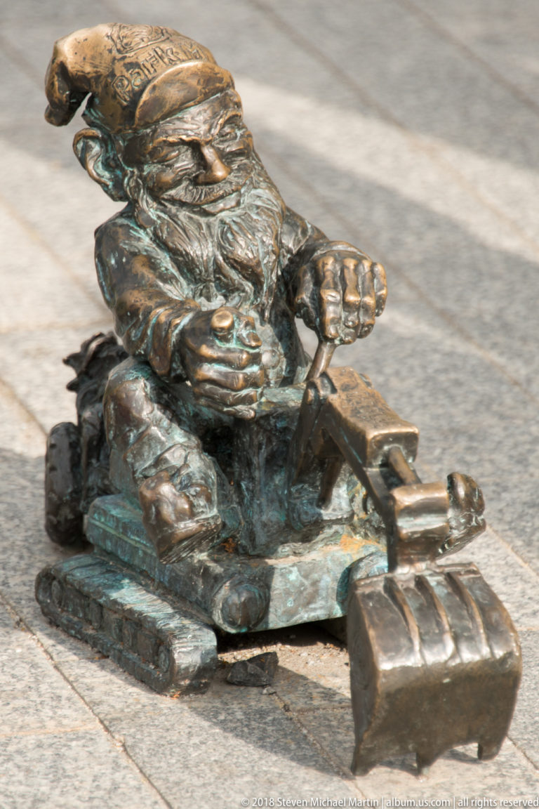 Bronze Wrocław dwarf seated at the controls of a miniature excavator with bucket extended.