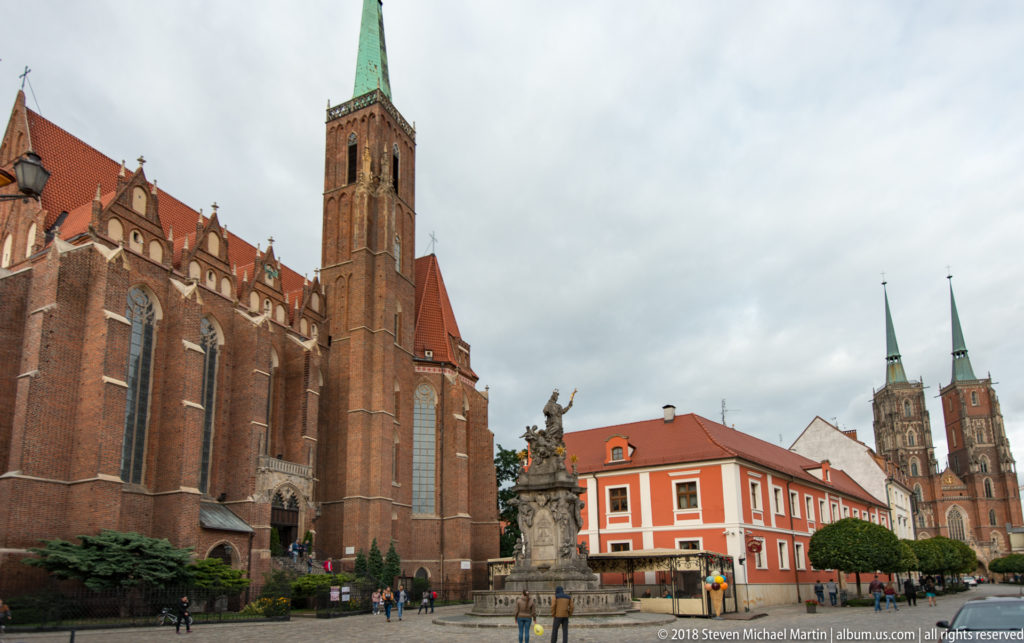 Large red-brick Gothic church in foreground with stone monument and Wrocław Cathedral’s twin towers in the distance.