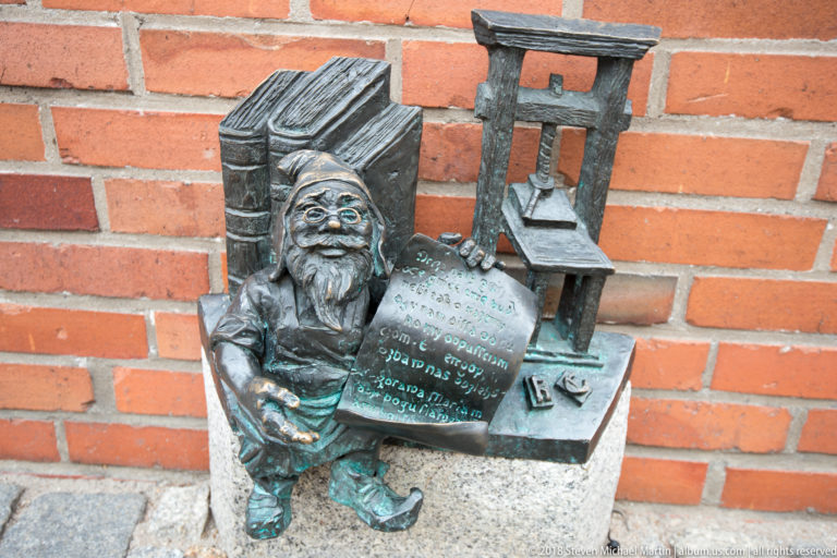 Bronze bearded dwarf sits beside miniature equipment and a small inscribed plaque against a red-brick wall.