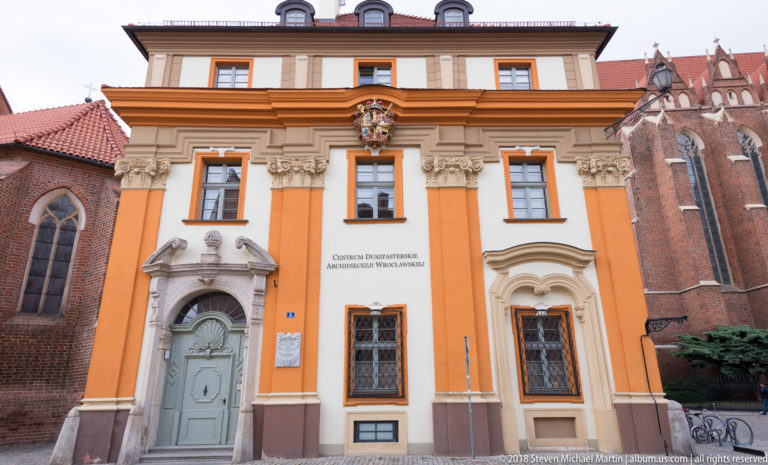 Orange-and-white Baroque-style institutional façade with crest, decorative portals, and adjoining brick church.