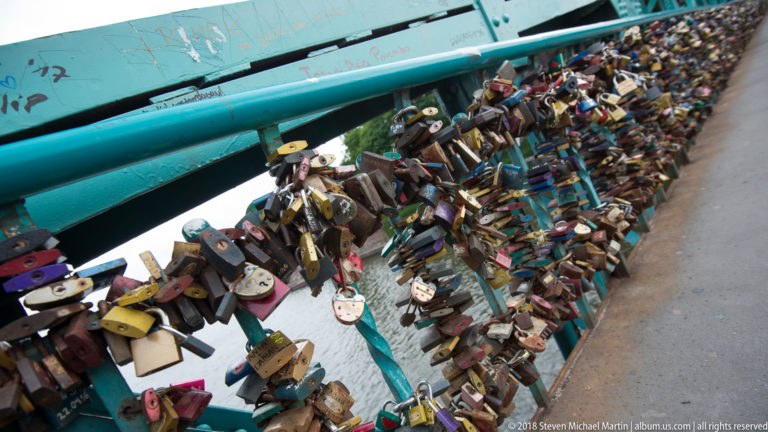 Large cluster of colorful padlocks densely attached to a turquoise-painted bridge railing.