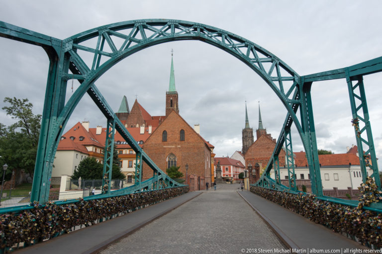 Turquoise iron bridge with padlock-covered railings framing church towers and red roofs beyond.