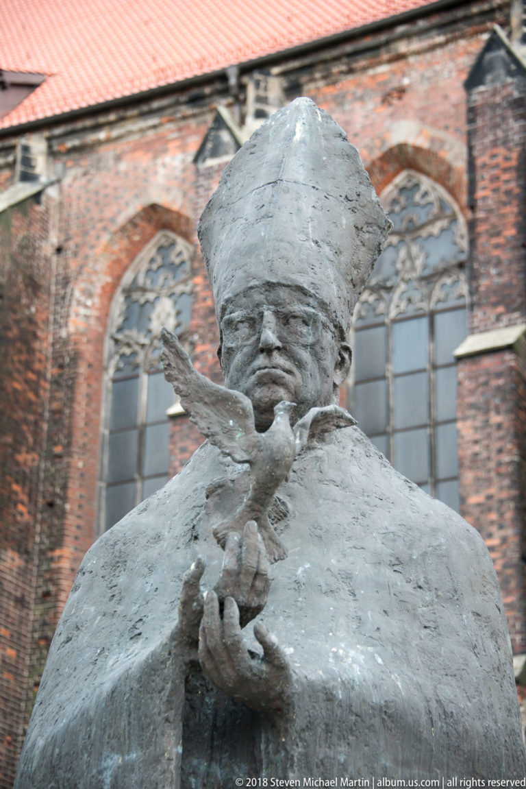 Weathered bishop figure in mitre holding a bird, with Gothic brick church windows in the background.