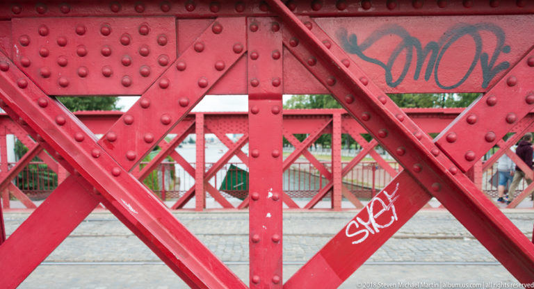 Close view of red riveted steel bridge trusses with diagonal braces and graffiti.