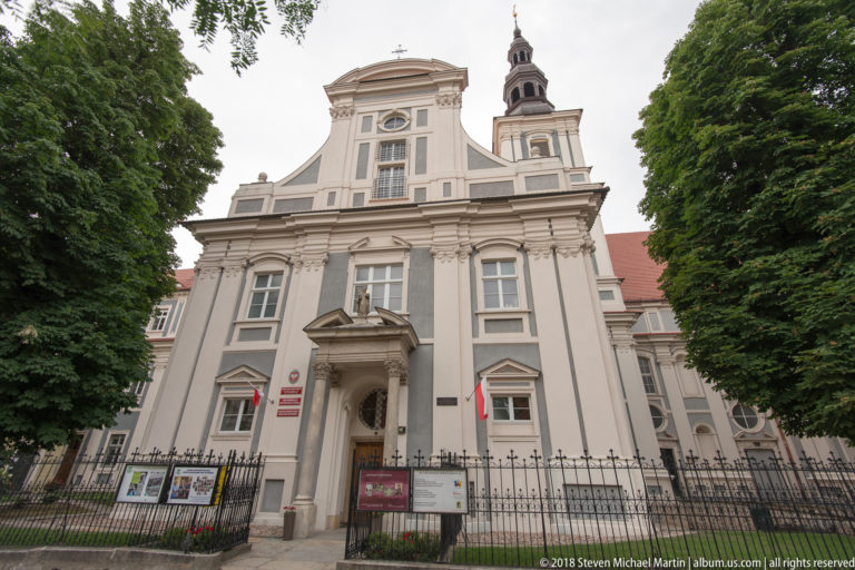 Baroque church façade with columned portal, tall pilasters, curving gable, bell tower, and iron fence.