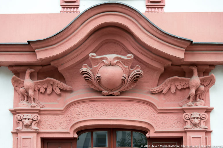 Salmon-colored ornamental window crest with central cartouche, winged creatures, scrolls, and carved floral details.