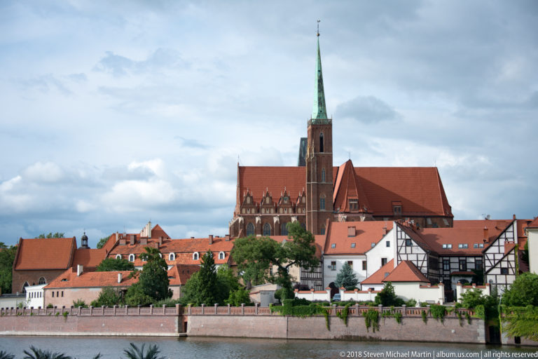 Riverside view of a tall Gothic church spire rising above red roofs and historic buildings in Wrocław.