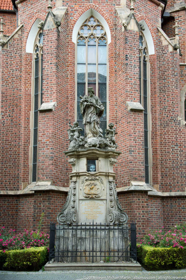 Ornate stone monument enclosed by iron railing in front of tall Gothic windows and red-brick church wall.