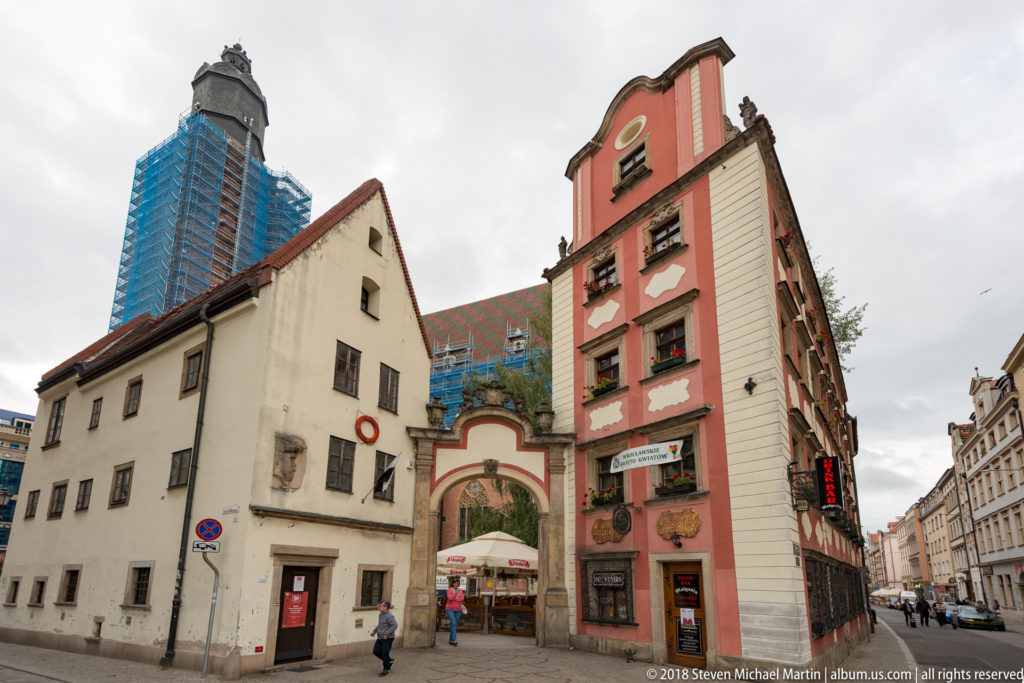 Narrow passage between pale and pink historic buildings, with painted archway and pedestrians at the entrance.