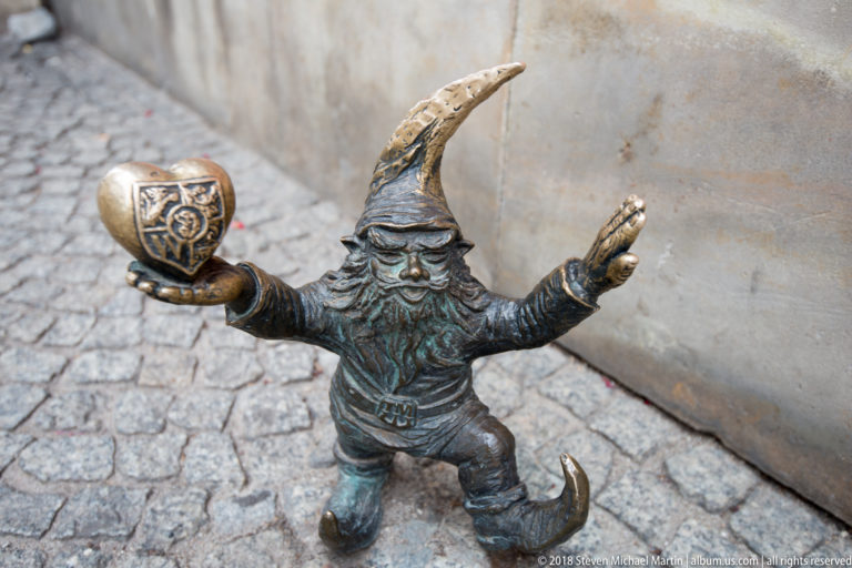 Bronze Wrocław dwarf in pointed hat holds a decorated heart while extending one arm.