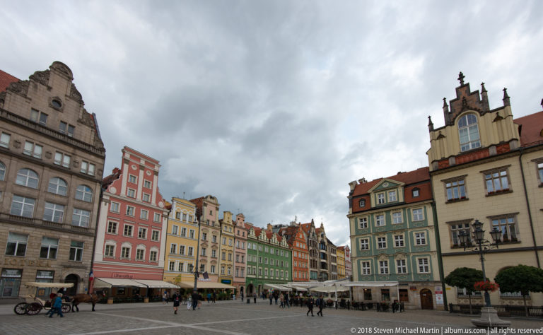 Wide market-square view with colorful historic façades, pedestrians, café seating, and cloudy sky.