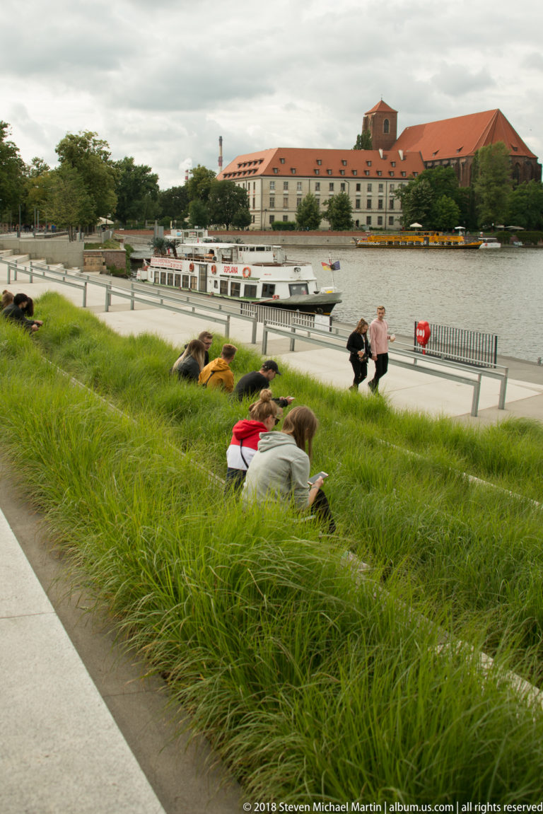 People lounge in tall green grass beside the river, with moored boats and large historic buildings across the water.