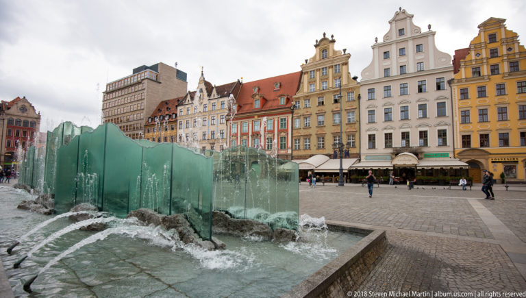 Green translucent glass fountain with churning water in foreground and colorful historic Rynek façades behind.