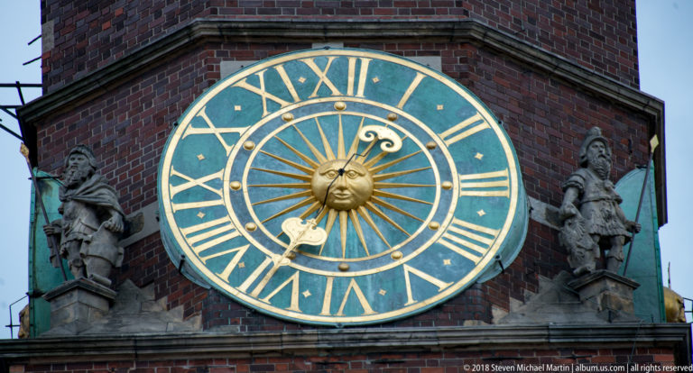 Large blue-green clock with gold Roman numerals and sun face, flanked by sculptures on a brick tower.