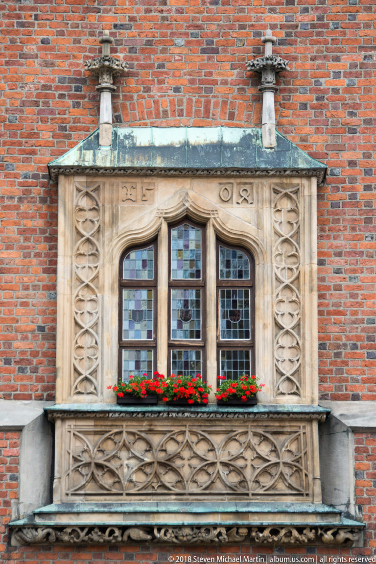 Carved stone window with leaded panes, geometric tracery, flower boxes, and red-brick wall at Wrocław Old Town Hall.