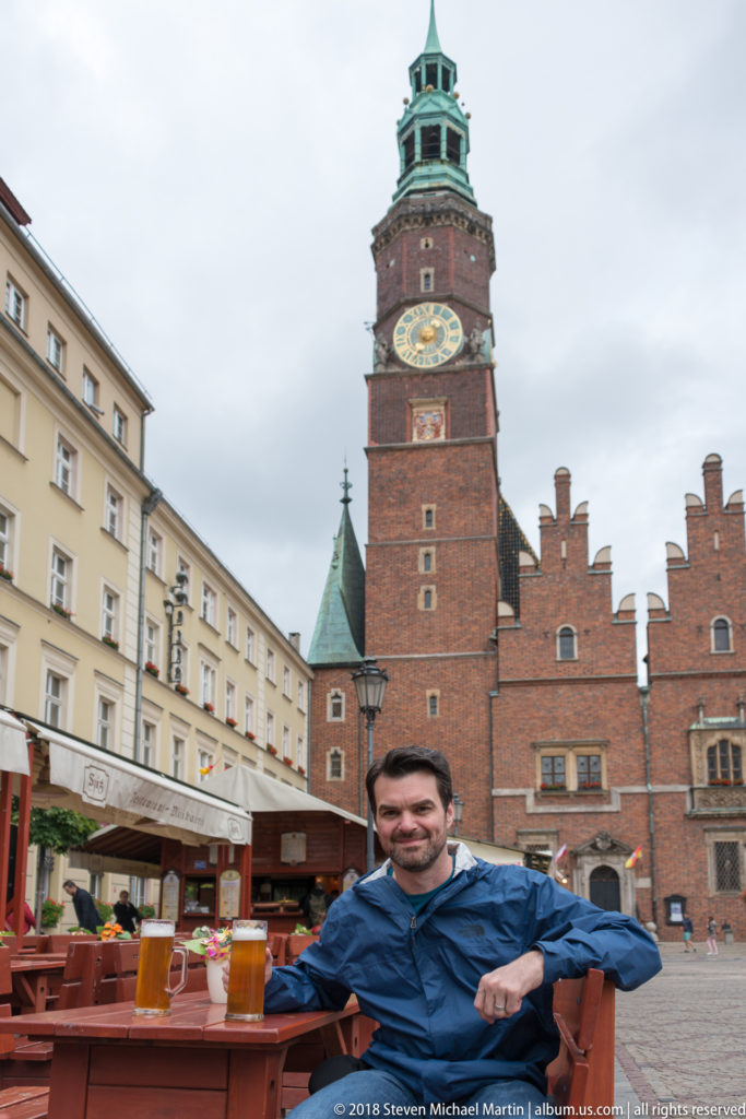 Seated visitor with beer at an outdoor café, with Wrocław Old Town Hall and clock tower behind.