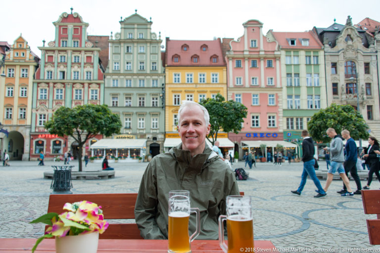 Smiling visitor seated at an outdoor table with two beers, colorful Wrocław market-square buildings behind.