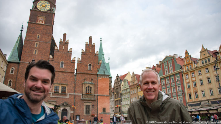 Two smiling visitors pose before Wrocław Old Town Hall and colorful historic façades in the market square.