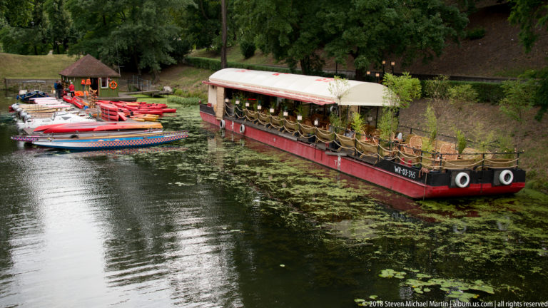 Covered red-and-cream passenger boat moored beside colorful paddleboats on a tree-lined waterway.