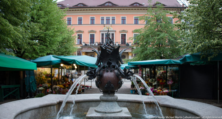 Dark ornamental fountain with arcing water jets, surrounded by flower stalls and a pastel historic building.