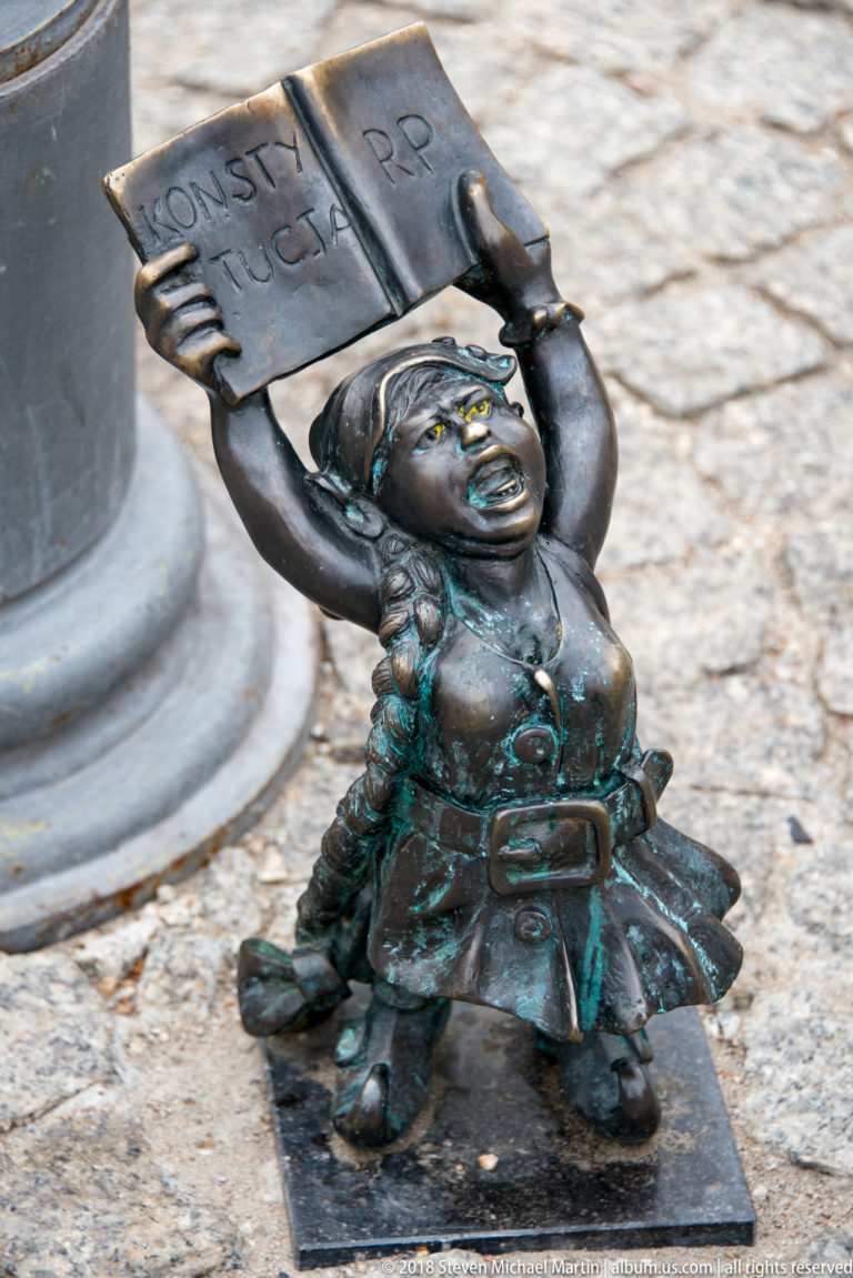 Bronze Wrocław dwarf seated on a small base while holding a rectangular plaque overhead.