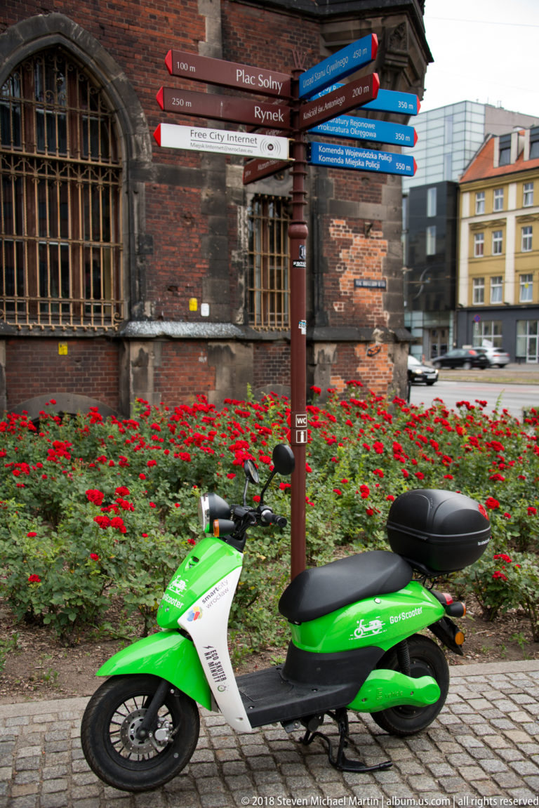 Green shared scooter parked below a directional signpost beside red flowers and an old brick building.
