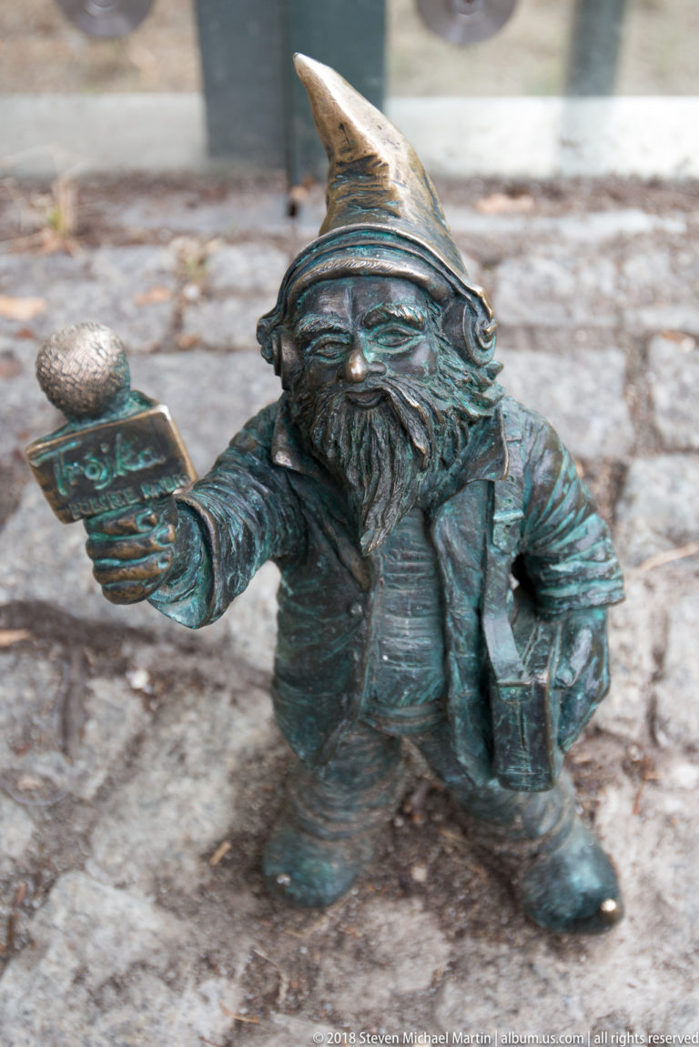 Bronze Wrocław dwarf in pointed hat extends one hand while standing on gray paving stones.