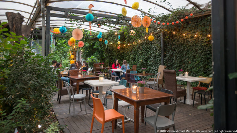 Courtyard café with wooden tables, colorful chairs, greenery, hanging decorations, and a transparent overhead canopy.