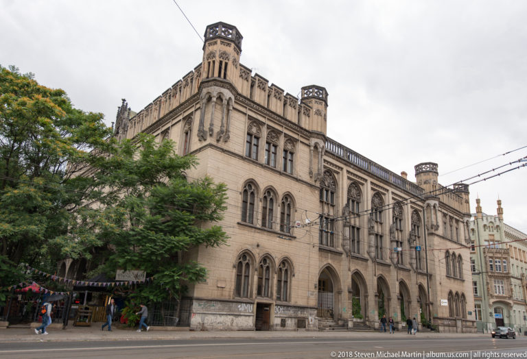 Large pale historicist building with arcades, crenellated roofline, corner towers, trees, and tram wires.
