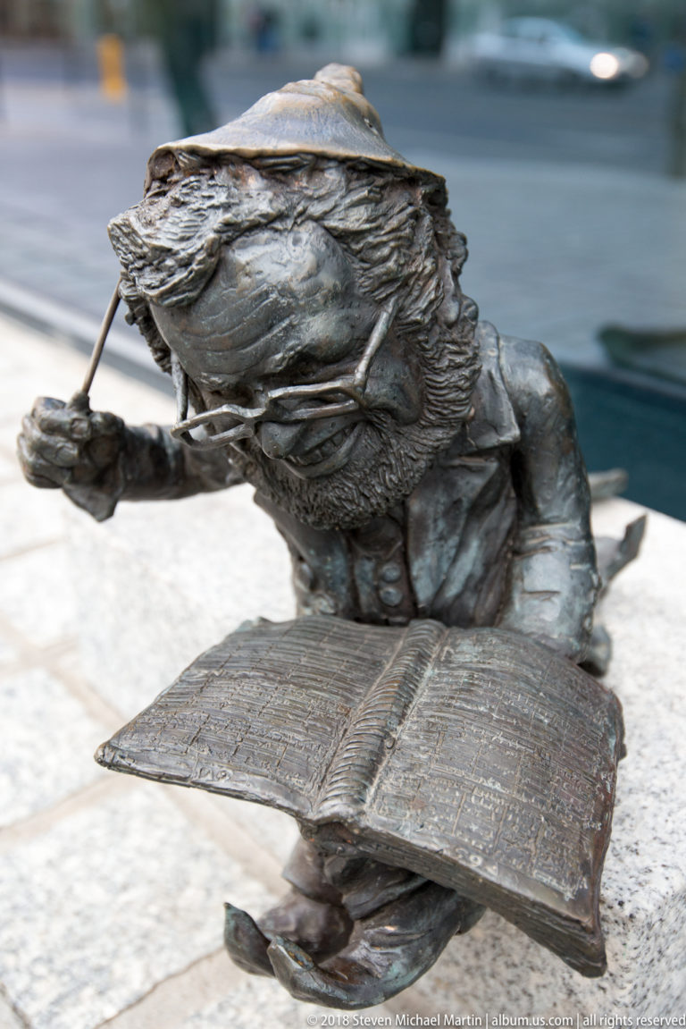Bronze Wrocław dwarf leans over an open engraved book in a small street-level sculpture.