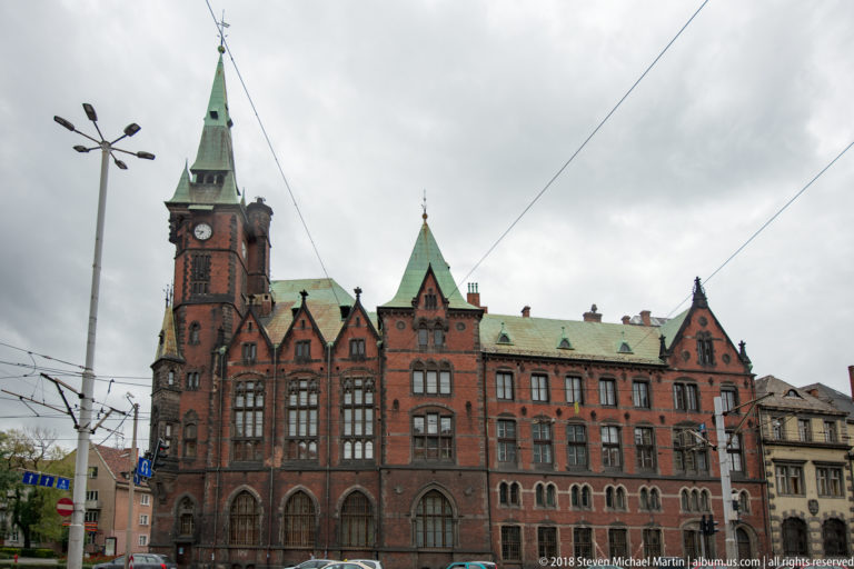 Large red-brick Neo-Gothic building with central clock tower, pointed gables, arched windows, and tram wires.