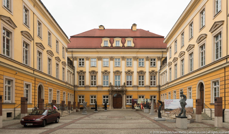 Symmetrical pale yellow courtyard with classical façades, central doorway, red roof, parked car, and sculpture.