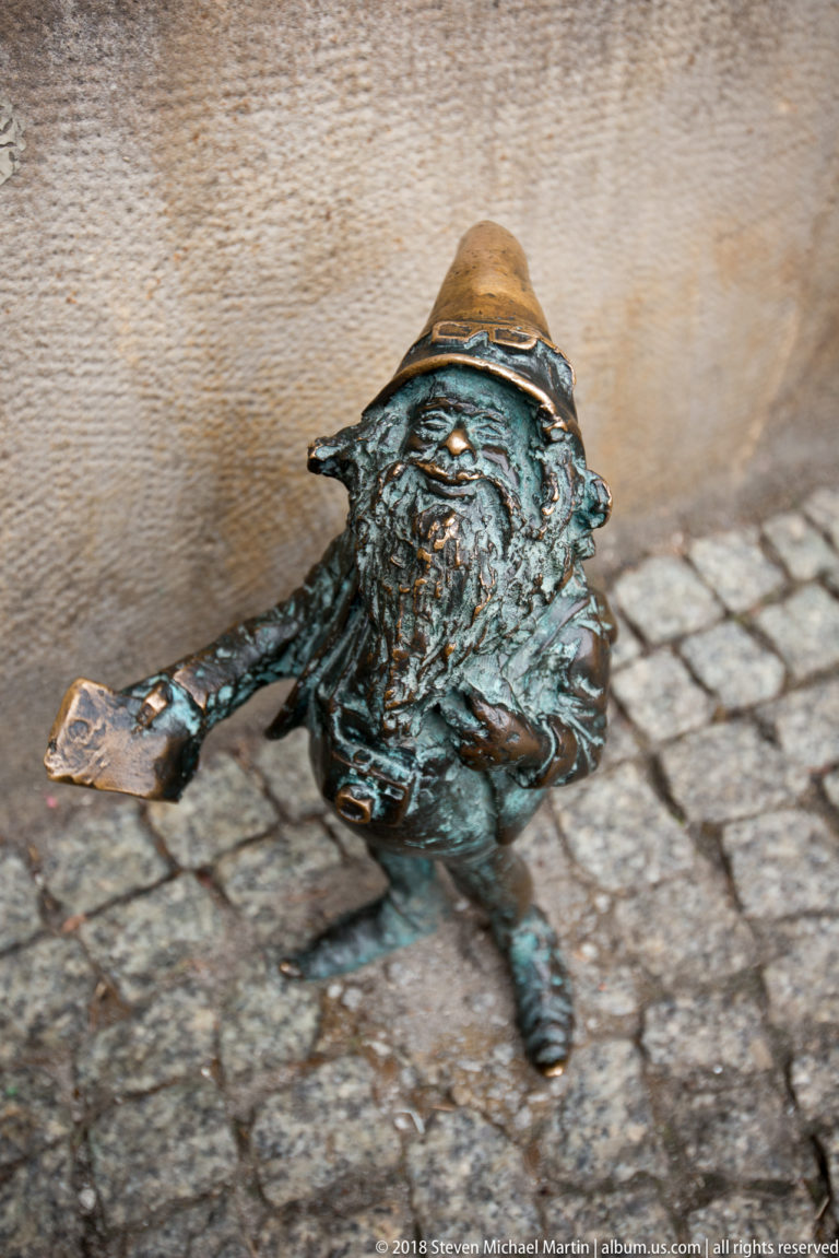 Small bronze Wrocław dwarf reclines beside a wall on cobblestone paving, wearing a broad hat.