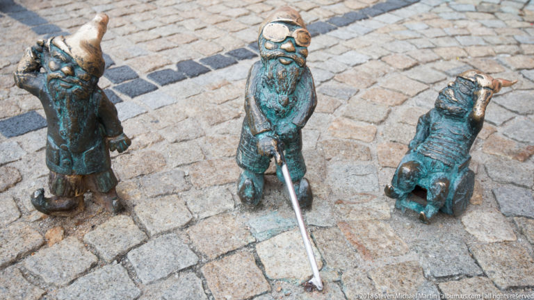Three small bronze Wrocław dwarf figures stand together on cobblestones, one holding a cane.