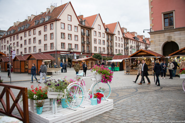 Decorative flower-filled bicycle in foreground with pedestrians, market stalls, and colorful historic buildings beyond.