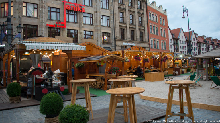 Wooden café stalls and standing tables occupy a paved historic square lined with narrow multistory façades.