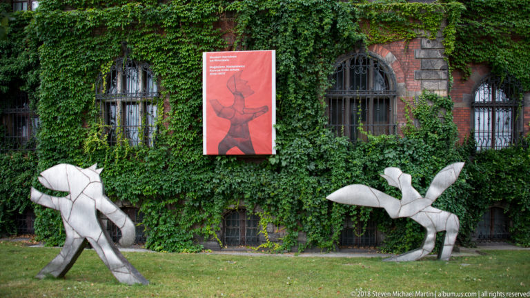 Two abstract pale metal sculptures on grass in front of an ivy-covered brick building and red banner.
