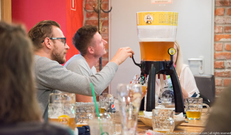 Tall Bierhalle tabletop beer dispenser surrounded by patrons, glasses, and mugs at a crowded wooden table.