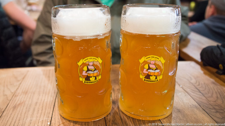 Two branded Bierhalle glasses filled with hazy golden wheat beer and thick foam on a wooden table.