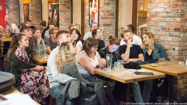 Crowded brick-walled beer hall with patrons at wooden tables watching a televised football match.