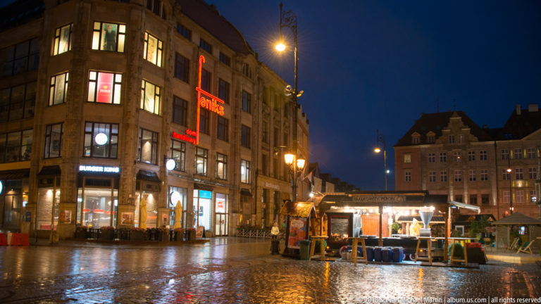 Temporary market stalls and illuminated storefronts reflected on wet paving in Wrocław Market Square at night.
