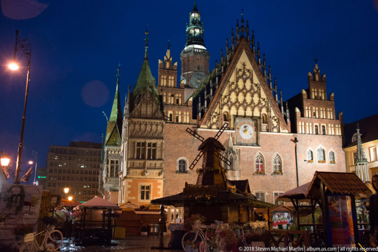 Illuminated east façade of Wrocław Old Town Hall with Gothic tracery, astronomical clock, and market stalls.